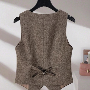 May include: A brown tweed vest featuring a sleeveless design, a V-neckline, and a decorative adjustable strap with a metal buckle at the lower back. The garment has a textured, speckled fabric finish and a pointed hemline at the bottom center.