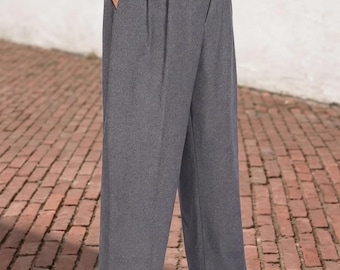 Könnte beinhalten: Graue Hose mit hochsitzendem Bund, doppelten Falten an der Vorderseite, Seitentaschen und umgeschlagenen Säumen. Die Hose wird mit einem braunen Ledergürtel mit goldfarbener Metallschnalle über einem hellen Hemd getragen.