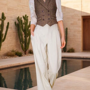 May include: A brown tweed double-breasted vest with dark buttons and notched lapels, worn over a white long-sleeved collared shirt paired with high-waisted, wide-leg cream trousers featuring front pleats and side pockets.