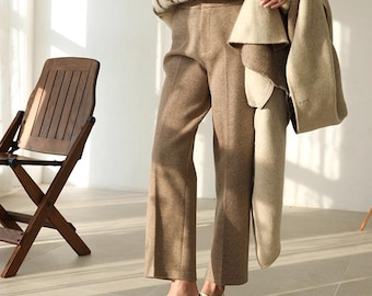 May include: Mid-rise trousers in a solid tan wool-like fabric featuring a straight-leg cut and a clean waistband. The trousers are paired with a cream-colored coat draped over the arm and white slip-on loafers with a low heel.