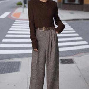 May include: A dark brown long-sleeved knit sweater paired with high-waisted, wide-leg gray tweed trousers. The outfit includes a thin brown leather belt with a gold-toned buckle and a delicate gold necklace with a small pendant.