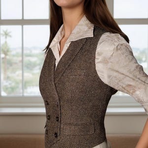 May include: A brown and black herringbone patterned double-breasted vest with a notched lapel, featuring four dark buttons on the front and two welt pockets, worn over a cream-colored long-sleeved shirt with a subtle tonal floral pattern.