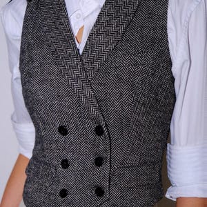 May include: A double-breasted vest featuring a black and white herringbone pattern, a notched lapel, and four black buttons. The vest is worn over a crisp white collared shirt with long sleeves and light-colored trousers.