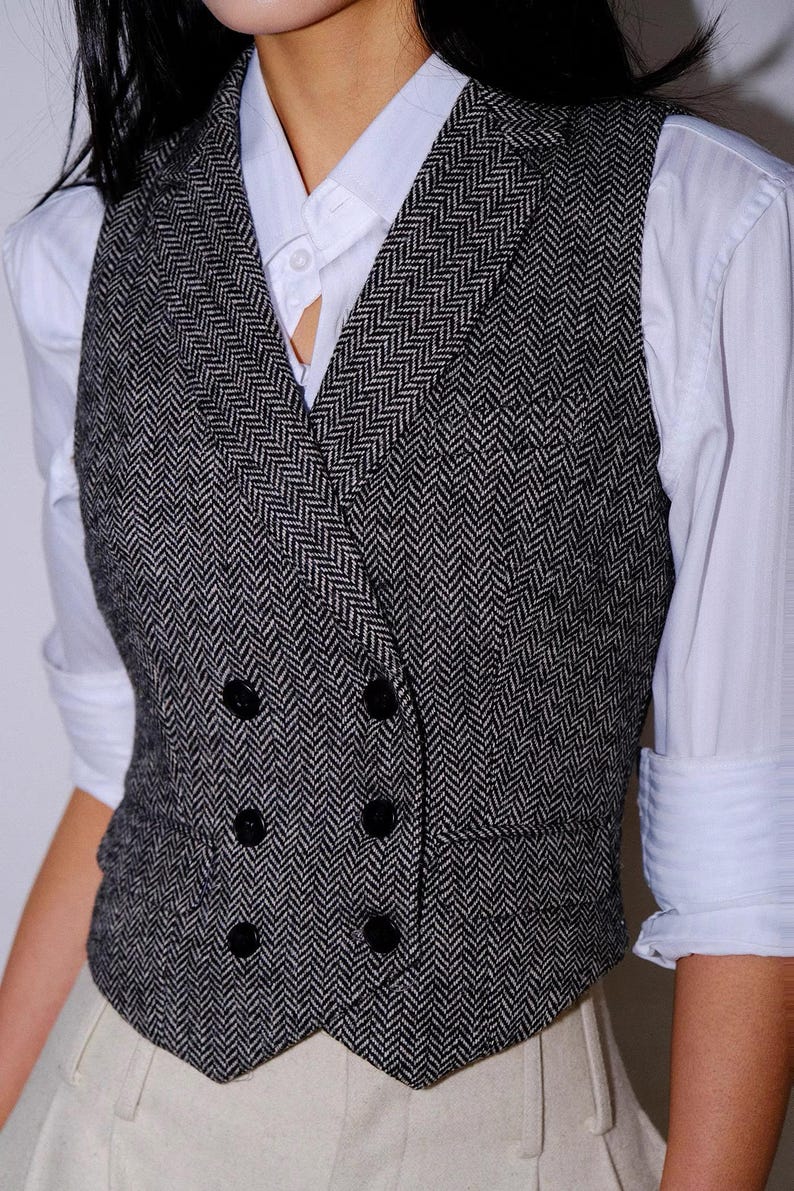 May include: A double-breasted vest featuring a black and white herringbone pattern, a notched lapel, and four black buttons. The vest is worn over a crisp white collared shirt with long sleeves and light-colored trousers.