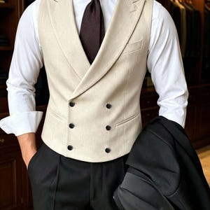 May include: A double-breasted beige vest with wide lapels and six black buttons, worn over a crisp white long-sleeved dress shirt with a dark brown necktie. The ensemble includes black trousers and a black suit jacket held in one hand.