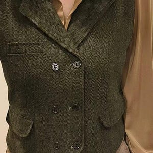 May include: A dark green double-breasted vest featuring a herringbone pattern, a notched lapel, and six black buttons. The vest includes two front flap pockets and one chest pocket. The text "Customer review photo - Deep Green Color" is visible.