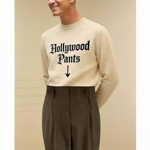 Peut inclure: Une personne porte un pull crème avec l'inscription "Hollywood Pants" en noir. Le pull est associé à un pantalon plissé marron foncé. La flèche pointe vers le bas, vers le pantalon.