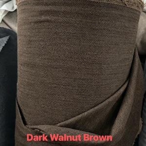 May include: A roll of dark walnut brown fabric featuring a woven herringbone pattern. The textile has a visible frayed top edge and includes printed text stating Dark Walnut Brown and About 450gsm on the lower portion of the material.