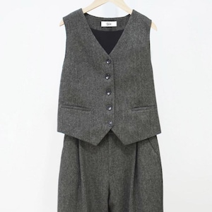 Peut inclure: Un gilet et un pantalon assortis en tissu gris foncé à motif chevrons. Le gilet présente une encolure en V, cinq boutons foncés et deux poches passepoilées. Une étiquette blanche avec le texte noir Vex est cousue à l'intérieur du col.