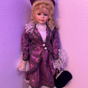 May include: A porcelain doll in Victorian attire. The doll is wearing a purple floral dress with white lace, a matching hat, and a black purse. The doll has blonde curly hair and blue eyes. The doll is approximately 40 cm tall.
