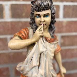 Puede incluir: Una figura de cerámica pintada de una persona joven con cabello oscuro recogido en un moño y ojos de color azul brillante. La figura viste una prenda de color claro con detalles en naranja y sostiene un trozo de tela frente a la boca.