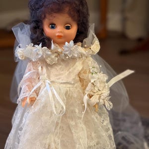 May include: A vintage doll in a white lace wedding dress and veil. The doll has dark hair, blue eyes, and a floral headpiece. It holds a small bouquet of white flowers and ribbons, a classic bridal theme.