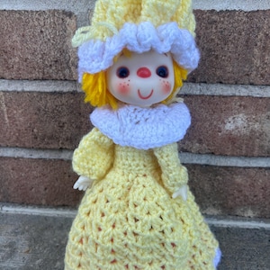 May include: A vintage doll with a painted face, yellow yarn hair, and a yellow crocheted dress. The doll has a white crocheted collar and hat with a ruffled edge. The doll's face has blue eyes, red freckles, and a red nose.