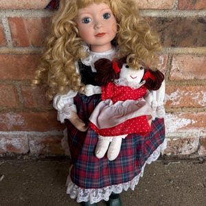 May include: A porcelain doll with blonde, curly hair, blue eyes, and a red tartan dress. The doll holds a smaller doll in a red polka dot dress. The doll's dress has white lace trim, and a matching tartan bow is in her hair.