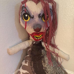 May include: A handmade doll with a scary clown-like face. The doll has red yarn hair, yellow eyes, and a red nose and mouth. The doll's body is a mix of brown and white patterned fabric, with white arms.