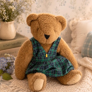 May include: A plush brown teddy bear with a soft, fuzzy texture, featuring a black stitched nose and closed eyes. The bear is wearing a sleeveless dress with a green and navy blue plaid pattern, accented by a small yellow detail at the centre.