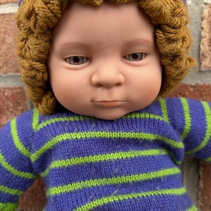 May include: A doll with a light skin tone, brown eyes, and a neutral expression. The doll wears a textured, mustard-brown crocheted hat and a long-sleeved, knitted jumper featuring horizontal stripes in alternating shades of purple and lime green.
