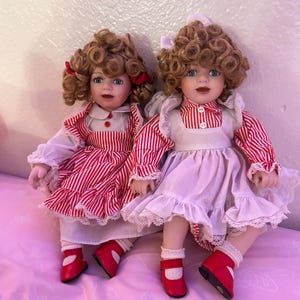 May include: Two porcelain dolls with curly brown hair and blue eyes. They wear red and white striped dresses, white aprons, and red shoes. Each doll has a white bow in their hair. The dolls are sitting on a pink floral patterned surface.