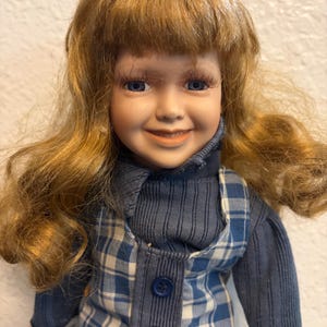 Peut inclure: Une poupée en porcelaine aux longs cheveux ondulés châtain clair, aux yeux bleus et aux longs cils. La poupée porte un haut à manches longues côtelé bleu foncé sous une robe chasuble à carreaux bleus et blancs avec trois boutons bleu foncé.