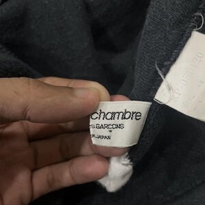Puede incluir: Primer plano de una prenda gris oscuro con una etiqueta blanca. La etiqueta dice "chambre des garcons" y "MADE IN JAPAN". Tambi&eacute;n es visible una segunda etiqueta con texto en japon&eacute;s.