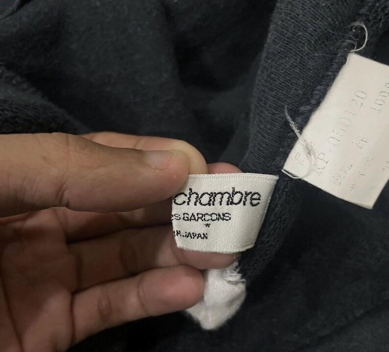 Puede incluir: Primer plano de una prenda gris oscuro con una etiqueta blanca. La etiqueta dice "chambre des garcons" y "MADE IN JAPAN". Tambi&eacute;n es visible una segunda etiqueta con texto en japon&eacute;s.