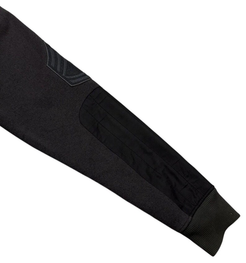 May include: Close-up of a dark grey sleeve with black patches and a ribbed cuff. The sleeve features a textured knit fabric and a patch on the upper arm with a subtle design. The black patches provide contrast.