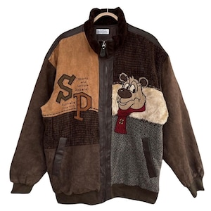 Op de afbeelding: Bruine en tan vintage jas met een cartoon berenpatch en de letters "SP" op de voorkant. De jas heeft een ritssluiting, een kraag van imitatiebont en een mix van texturen, waaronder suède en een visgraatpatroon.