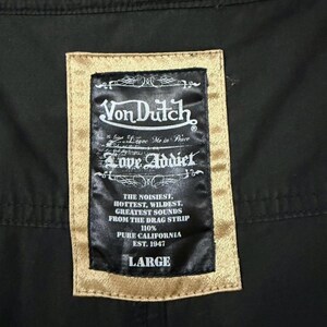 Puede incluir: Primer plano de una tela negra con una etiqueta rectangular. La etiqueta tiene un borde dorado y presenta el logotipo "Von Dutch", las palabras "Love Addict" y otro texto. El tama&ntilde;o "LARGE" est&aacute; impreso en la parte inferior.