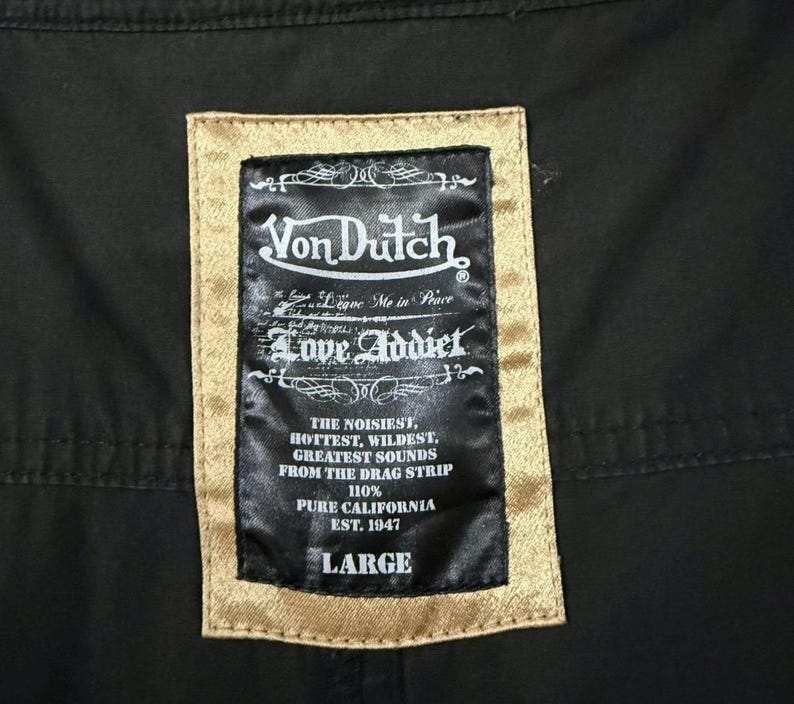 Puede incluir: Primer plano de una tela negra con una etiqueta rectangular. La etiqueta tiene un borde dorado y presenta el logotipo "Von Dutch", las palabras "Love Addict" y otro texto. El tama&ntilde;o "LARGE" est&aacute; impreso en la parte inferior.