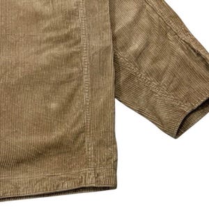 K&ouml;nnte beinhalten: Nahaufnahme einer beigefarbenen Cordjacke. Der Stoff hat eine gerippte Textur mit vertikalen Linien, die das Material hinunterlaufen. Der &Auml;rmel ist sichtbar und zeigt die Manschette und die Nahtdetails.