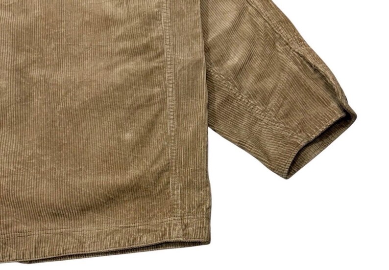 K&ouml;nnte beinhalten: Nahaufnahme einer beigefarbenen Cordjacke. Der Stoff hat eine gerippte Textur mit vertikalen Linien, die das Material hinunterlaufen. Der &Auml;rmel ist sichtbar und zeigt die Manschette und die Nahtdetails.