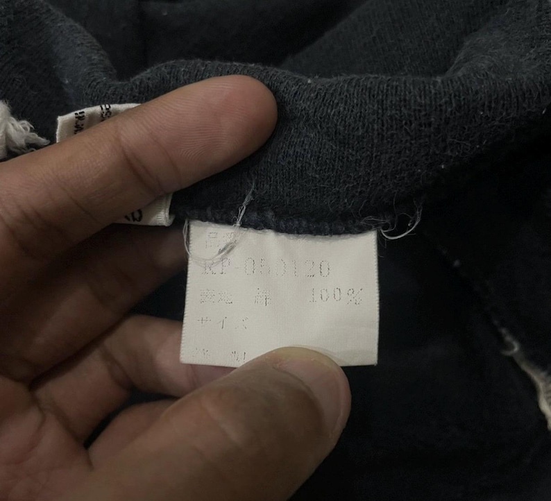 Puede incluir: Primer plano de una tela gris oscuro con una etiqueta blanca. La etiqueta tiene texto japon&eacute;s y el n&uacute;mero "RP-050120". La tela parece ser un material suave, posiblemente algod&oacute;n o una mezcla.