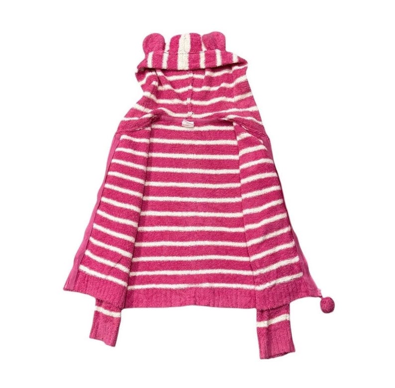 May include: A pink and white striped hooded sweater with a cat ear design on the hood. The sweater has long sleeves and a pom-pom detail at the end of each sleeve. The sweater is open in the front.