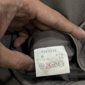 May include: A close-up of a clothing tag with the number X560222 printed on it. The tag also includes Japanese characters and the percentages 95% and 5%. The tag is attached to a gray fabric.