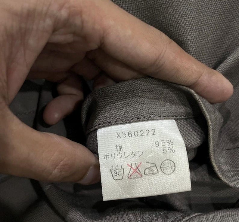 May include: A close-up of a clothing tag with the number X560222 printed on it. The tag also includes Japanese characters and the percentages 95% and 5%. The tag is attached to a gray fabric.