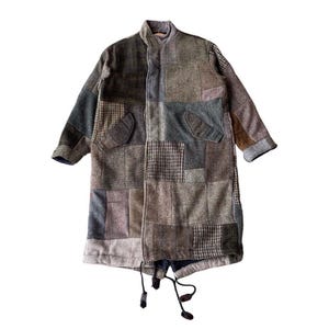 Puede incluir: Abrigo largo de patchwork en tonos marrones, grises y verdes. El abrigo tiene cuello alzado, dos bolsillos delanteros y un cordón en el dobladillo. Está confeccionado con diferentes tejidos texturizados.