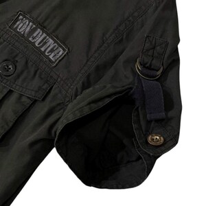 Puede incluir: Primer plano de una camisa verde oscuro con el texto "Von Dutch" en un parche. La camisa tiene bolsillos abotonados, una manga abotonada y una correa azul oscuro con un anillo de metal.
