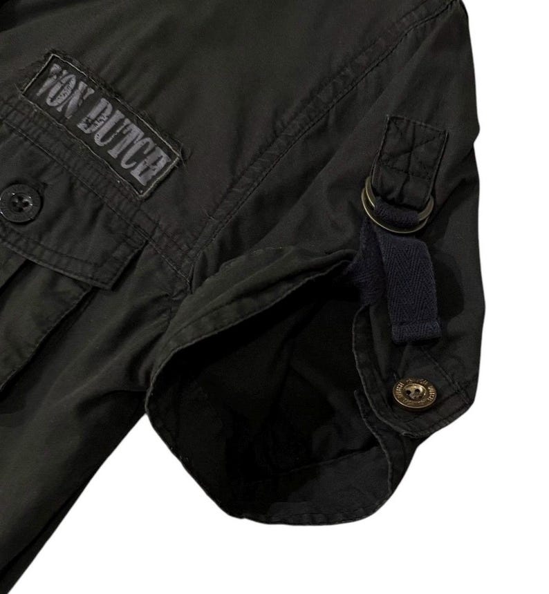 Puede incluir: Primer plano de una camisa verde oscuro con el texto "Von Dutch" en un parche. La camisa tiene bolsillos abotonados, una manga abotonada y una correa azul oscuro con un anillo de metal.