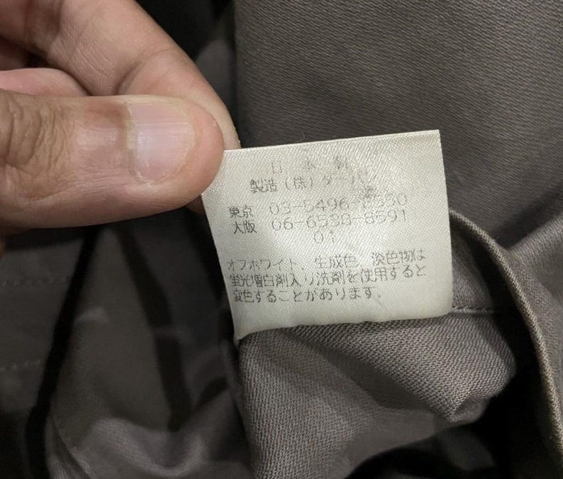 May include: Close-up of a tag with Japanese text and numbers. The tag is white with black text and is attached to a gray fabric. The text includes the words "Tokyo" and "Osaka" along with phone numbers.
