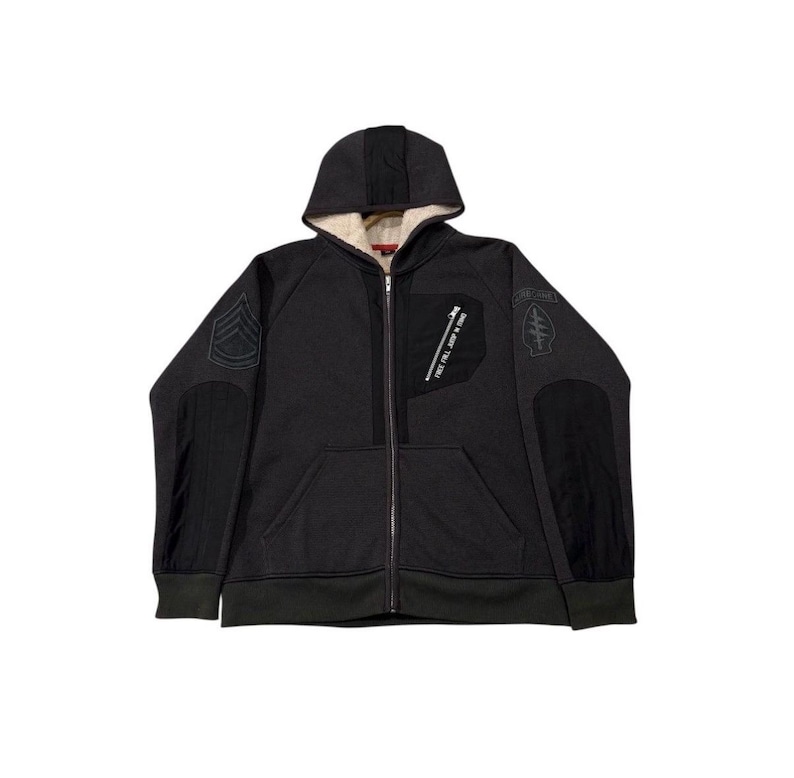 May include: Dark grey zip-up hoodie with a hood and a cream-coloured lining. The jacket features black patches on the sleeves and chest, with text and military-style insignia. The hoodie has a full-length zip and ribbed hem and cuffs.