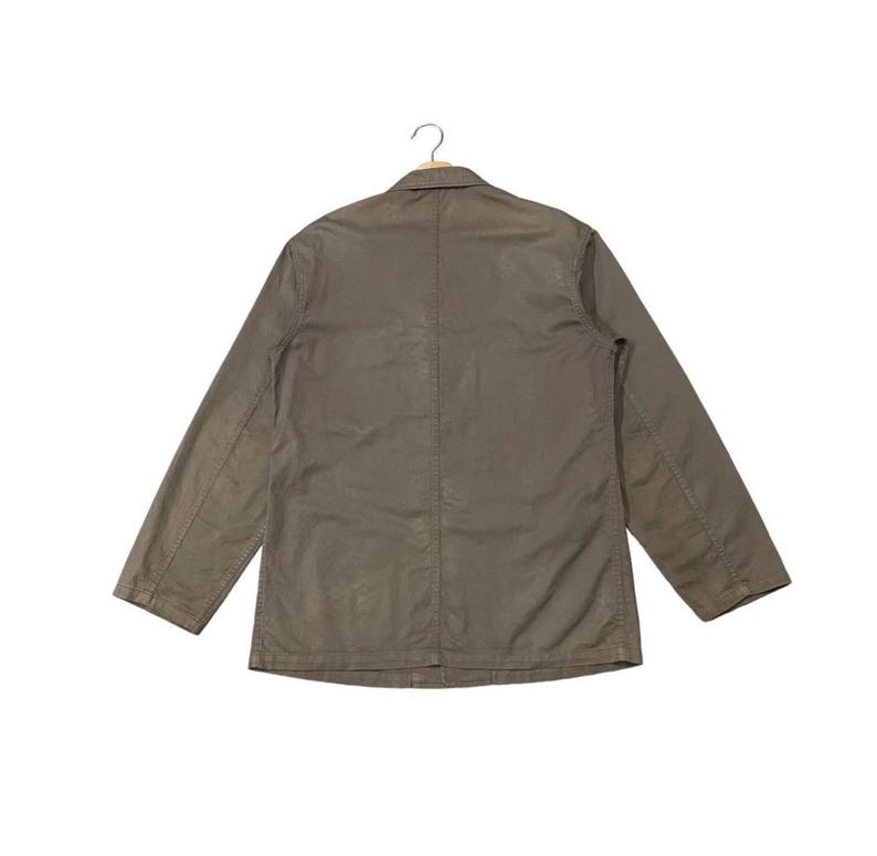 May include: A khaki-colored jacket with long sleeves and a button closure. The jacket is hanging on a hanger against a white background. The jacket has a simple, classic design.