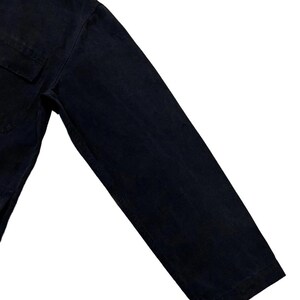 May include: Close-up of a black long-sleeved shirt. The shirt has a single chest pocket and a buttoned cuff. The fabric appears to be a sturdy, dark-coloured material.