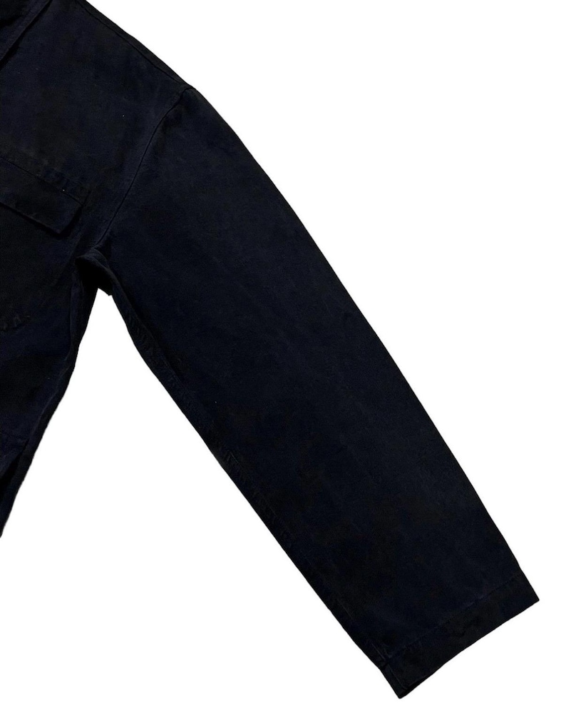 May include: Close-up of a black long-sleeved shirt. The shirt has a single chest pocket and a buttoned cuff. The fabric appears to be a sturdy, dark-coloured material.