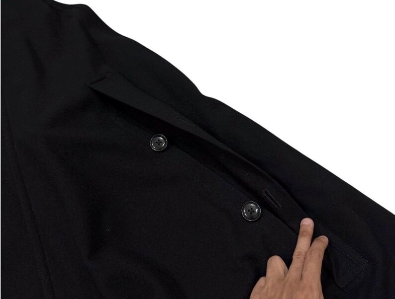 May include: Close-up of a black coat with two visible buttons and a pocket. The coat is made of a dark fabric and has a classic design. The buttons are round and black, and the pocket is rectangular.