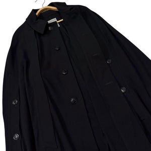 May include: A black coat with a classic design, featuring a collar, button closures, and a tailored fit. The coat is hanging on a wooden hanger, showcasing its structure and style. The coat's dark color and simple design suggest a versatile garment.
