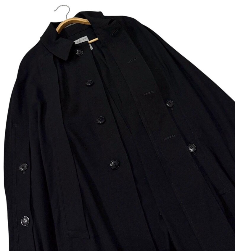 May include: A black coat with a classic design, featuring a collar, button closures, and a tailored fit. The coat is hanging on a wooden hanger, showcasing its structure and style. The coat's dark color and simple design suggest a versatile garment.