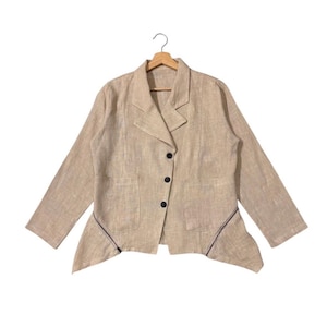 Puede incluir: Blazer beige con solapa de muesca, dos bolsillos delanteros y tres botones negros. La chaqueta de manga larga tiene un dobladillo asimétrico con ribete oscuro y está colgada de una percha de madera.