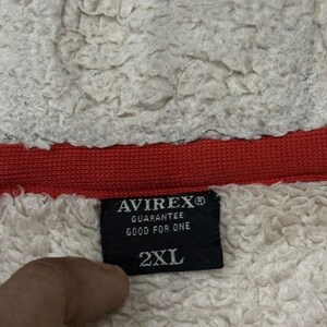 May include: A close-up shot of a black label with the brand name "AVIREX" and the text "GUARANTEE GOOD FOR ONE" in white. The label also indicates a size of "2XL". The label is set against a backdrop of white, textured fabric and a red band.