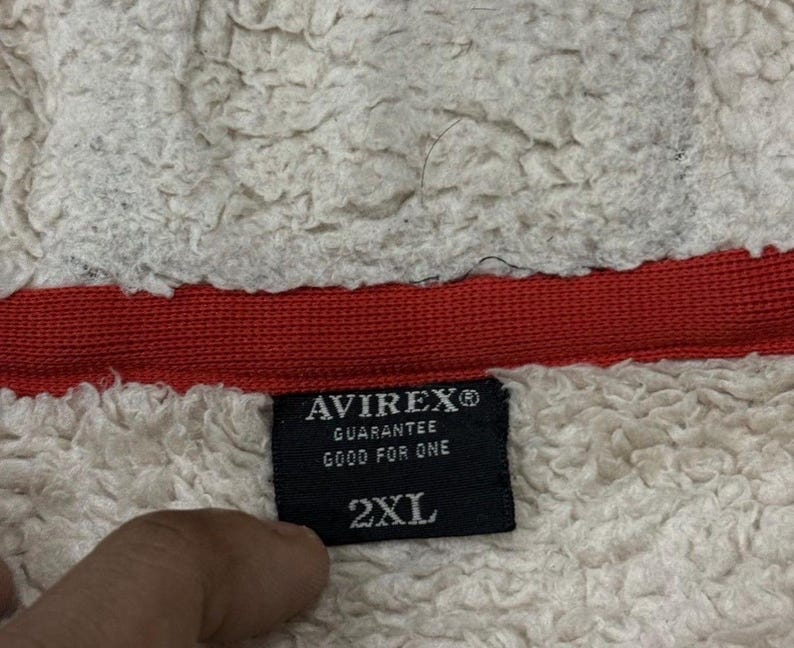 May include: A close-up shot of a black label with the brand name "AVIREX" and the text "GUARANTEE GOOD FOR ONE" in white. The label also indicates a size of "2XL". The label is set against a backdrop of white, textured fabric and a red band.
