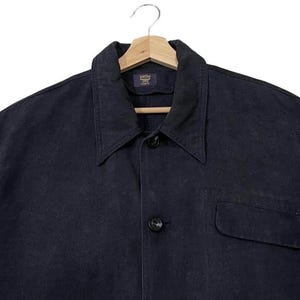 May include: A dark navy blue shirt with a collar and a single pocket, hanging on a wooden hanger. The fabric looks like cotton or denim. The shirt has a classic design. The buttons are black. The shirt is a versatile piece of clothing.